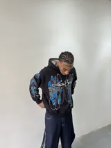 Hoodie oversize de color negro con estampado artístico en pecho y mangas en tonos azul y gris. Presenta capucha forrada con tejido a cuadros, costuras reforzadas y grifa de la marca en la manga.