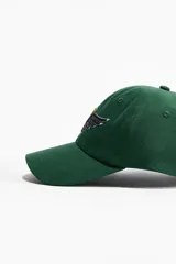 Gorra de sarga de algodón color verde, con diseño de seis paneles y visera curva. Presenta un bordado frontal con la letra A en color dorado y un ala en color azul oscuro. Incluye cinta antitranspirante interna y cierre metálico ajustable en la parte posterior.