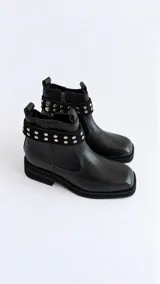 Botas cortas de cuero negro, con punta cuadrada, suela plana y taco bajo. Presentan una tira desmontable alrededor del tobillo decorada con tachas metálicas plateadas y cierre lateral interno.