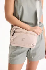 Bandolera color beige, con múltiples compartimentos frontales con cierre de cremallera, tiradores metálicos y correa ajustable para el hombro. Presenta un pequeño estampado con la inscripción 'everyday wear' en el frente.