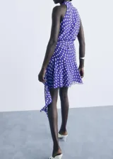 Vestido corto de poliéster con estampado de lunares, cuello en pico cruzado y diseño evasé. Presenta tirantes anchos y cierre mediante lazo lateral.