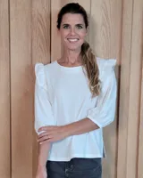 Blusa blanca de algodón con cuello redondo, mangas tres cuartos con puños elásticos y detalle de volados en los hombros.