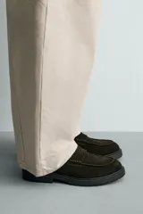 Mocasines de vestir color marrón oscuro, confeccionados en piel con acabado en gamuja serraje. Presentan detalle de antifaz en el empeine con costuras pespunteadas, punta redonda y suela track de goma robusta.