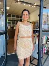 Conjunto de dos piezas color beige claro, compuesto por un top corto de tirantes finos y una falda corta. Ambas prendas están decoradas con flecos colgantes dispuestos en patrón de rombos sobre la tela base.