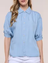 Camisa de tencel liviano estilo denim, color celeste, con cuello camisero, cierre frontal con botones nacarados, mangas cortas abullonadas con puños elásticos en punto smock y detalles de ojalillos.