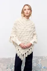 Poncho de tejido de punto grueso color crudo con diseño de trenzas, cuello alto y terminación con flecos en el ruedo.