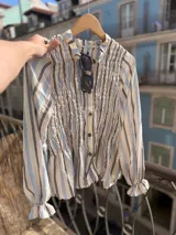Camisa de manga larga con diseño a rayas verticales en tonos azul, gris y crema. Presenta un cuerpo con frunces elásticos decorativos, cuello con volados y puños con elástico y terminación en volado. Incluye cierre frontal con botones.