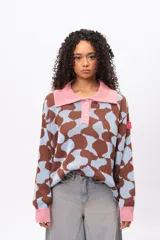 Buzo de punto con diseño geométrico abstracto en tonos marrón y celeste. Presenta cuello tipo polo en color rosado con cierre frontal de botones, mangas largas con puños rosados y un pequeño parche con logo en la manga izquierda.