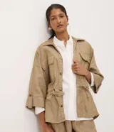 Campera tipo parka de la marca Marfinno, confeccionada en mezcla de algodón, viscosa y lino en color beige. Presenta cuello camisero, cierre frontal con botones, bolsillos con solapa en el pecho y cintura ajustable con cordón y lazo. Las mangas son largas con puños abotonados.