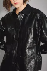 Campera de cuero sintético negro con acabado brillante, diseño tipo camisa con cuello clásico, cierre frontal con botones a presión y bolsillos delanteros. Cuenta con forro interior de algodón.