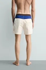 Short de baño blanco marfil con cintura marrón y cordón ajustable del mismo color. Presenta franjas laterales en tono azul marino con finos bordes rojizos y bolsillos laterales.