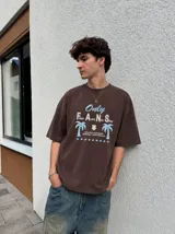 Remera oversize marrón con estampado gráfico frontal en tonos celestes y blancos con la leyenda "Only Fans", palmeras y estrellas.