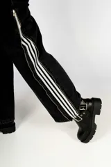 Pantalón de jean negro con corte recto, tiro alto, bolsillos delanteros y traseros, cierre con cremallera y botón. Presenta tres líneas blancas verticales a lo largo de la pierna y cierres metálicos dorados.