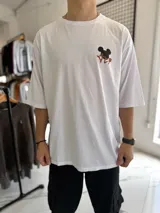 Remera oversize blanca de manga corta, con cuello redondo y etiqueta decorativa en la parte posterior del cuello. Presenta un gran estampado en la espalda que combina un diseño estilizado de Mickey Mouse con caracteres en estilo asiático en color rojo.