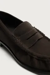 Mocasín de vestir para hombre confeccionado en piel con acabado en gamuza, color marrón. Presenta diseño tipo penny loafer con detalle de antifaz en el empeine, costuras visibles y suela contrastante con un ligero tacón.