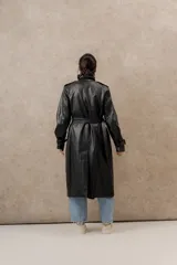 Trench largo de ecocuero negro, con cuello clásico, cierre cruzado con botones laterales y cinturón ajustable.