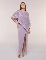 Vestido largo de seda fría color lila, de corte recto con tajo frontal, escote a la base y mangas tres cuartos. Cuenta con una capa superpuesta de encaje con bordado de lentejuelas y diseño asimétrico.