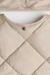 Campera acolchada de color beige con diseño guateado en rombos, hombros caídos, cuello redondo, cierre frontal de botones y bolsillos parche delanteros. Incluye una bufanda a juego que se abrocha en la nuca y cuenta con aislamiento térmico THERMOLITE.