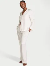 Pijama largo de satén color blanco marfil, compuesto por una camisa de manga larga con solapas, botones y bolsillo en el pecho, y un pantalón largo de corte recto con detalles de ribetes rosados. Presenta un sutil estampado de monograma jacquard en toda la prenda.