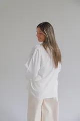 Camisa blanca de corte holgado con cuello clásico, cierre frontal de botones y mangas tres cuartos con sutil detalle bordado en los puños.