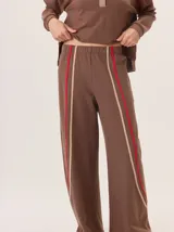Pantalón de corte ancho y estilo comfy, confeccionado en algodón color marrón. Presenta detalles de franjas verticales en tonos beige y rojo a lo largo de las piernas, con cintura elástica.