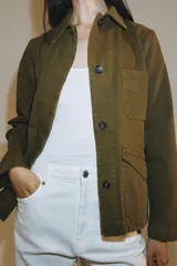 Chaqueta de estilo utilitario color verde militar, con cuello solapa, manga larga y cierre frontal con botones ocultos por solapa. Presenta bolsillos con solapa en la parte delantera y un bolsillo de plastrón en el pecho.