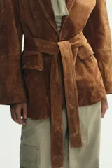 Chaqueta tipo blazer confeccionada en piel de ante, con cuello de solapa, hombreras y manga larga. Presenta cierre frontal con botones, bolsillos delanteros con solapa y un cinturón lazada del mismo material para ajustar a la cintura.