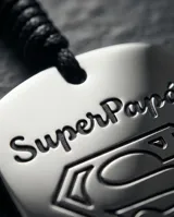 Llavero de acero inoxidable con placa cuadrada grabada con el logo de Superman y la frase Super Papá, unido a una correa trenzada de color negro y argolla metálica circular.