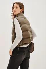 Chaleco acolchado de corte corto, color verde oliva, con cuello alto ajustable mediante cordones elásticos y cierre frontal con cremallera.