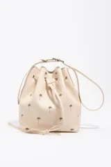 Bolso tipo saco color beige de piel vacuna, con estampado de palmeras pequeñas en marrón, cordón de ajuste frontal y asa regulable con hebilla metálica. Incluye el logo grabado de la marca en la parte inferior.