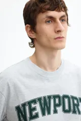 Remera de algodón gris jaspeado con cuello redondo y mangas cortas. Presenta un estampado frontal con la inscripción 'NEWPORT' en tipografía universitaria verde oscuro y 'RHODE ISLAND' debajo.