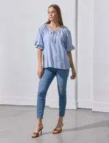 Blusa celeste y blanca a rayas verticales, de lino y corte holgado. Tiene cuello redondo con escote en V y lazo de ajuste. Las mangas son cortas y amplias, con volados en el borde.