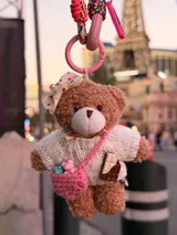 Llavero de peluche con forma de oso, vestido con un sweater tejido de punto color crema. El oso lleva un lazo con lunares en la cabeza, una pequeña cartera tejida color rosa con figuras de ositos en su interior y una pieza decorativa en forma de barra de chocolate.