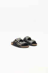 Sandalia plana negra de estilo 'dos hebillas' con tachas metálicas doradas decorando las tiras y hebillas ajustables doradas. La plantilla es de corcho y la suela de EVA.