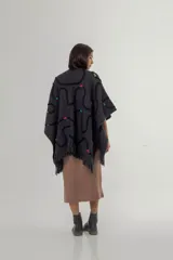 Ruana de tejido de punto grueso en color gris oscuro, con diseño de líneas curvas en relieve de color negro y detalles de costuras decorativas en colores vibrantes. Presenta terminaciones con flecos en el ruedo y una etiqueta de cuero sintético en la parte inferior.