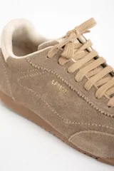 Zapatilla deportiva de gamuza color visón, con cordones a tono y suela de goma marrón. Presenta un diseño retro de running con el nombre de la marca grabado en dorado en el lateral.