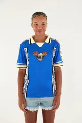 Camiseta deportiva Farm Etc. azul con cuello polo en tono crema y detalles negros, manga corta con líneas decorativas y estampado frontal con escudo y palmeras.