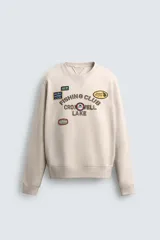 Sudadera de cuello redondo y manga larga en color beige. Presenta un diseño con texto bordado y múltiples parches decorativos en el frente, con terminaciones en rib en puños y cintura.