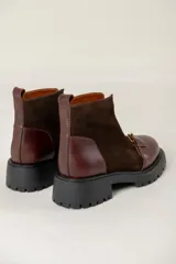 Bota de caña corta combinada en cuero liso y gamuza color marrón. Presenta cierre frontal con cremallera, tira con hebilla metálica decorativa en el empeine y suela track de goma negra.