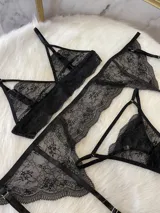 Conjunto de lencería de encaje negro compuesto por un bralette triangular, una tanga y un portaligas integrado. El diseño presenta detalles de encaje floral, breteles regulables y tiras elásticas ajustables.
