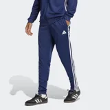 Pantalón deportivo Adidas Tiro 25 Essentials, color azul marino, con corte clásico y puños ajustados. Presenta las tres franjas características de Adidas en blanco a lo largo de las costuras laterales y el logo de la marca en blanco en el muslo.