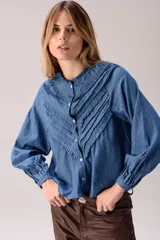 Camisa de denim azul con cuello mao y múltiples filas de volados en forma de V en el frente. Tiene mangas largas abullonadas con puños ajustados y cierre frontal de botones.