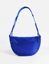 Bolso de hombro tipo media luna en color azul vibrante, confeccionado en material sintético. Presenta un diseño minimalista con cierre superior y correas laterales ajustables mediante cordones decorativos.