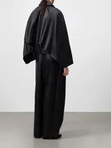 Chaqueta negra de piel ovina con diseño desestructurado, cuello solapa, cierre frontal con cremallera y bolsillos de vivo inclinados. Presenta una capelina en la espalda y mangas largas.