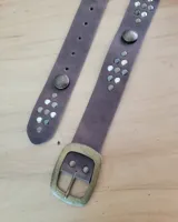 Cinto de cuero color marrón topo, con hebilla rectangular de metal envejecido y tachas redondas plateadas y oscuras a lo largo de la tira.