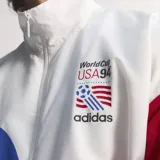 Campera deportiva Adidas de tejido técnico con cierre frontal y cuello alto. Presenta un diseño estampado con motivos de la Copa del Mundo USA 94, combinando colores blanco, azul y rojo. Cuenta con bolsillos laterales con cremallera y puños elásticos.