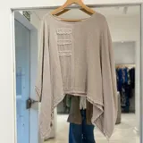 Blusa de lino color beige, de corte asimétrico y holgado, con mangas amplias, escote redondo y detalles de parches rectangulares deshilachados en el pecho.
