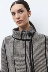 Capa de tejido de punto en color beige y negro con patrón de espiga, cuello alto y cierre de botón. Presenta vivos negros en contraste que delinean el diseño.