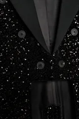 Blazer asimétrica negra de la línea ZW Collection, recubierta de lentejuelas tono sobre tono, con cuello solapa, manga larga y diseño cruzado con botones y cierre frontal con gancho metálico. Presenta bajo asimétrico con abertura y pliegues en la espalda.