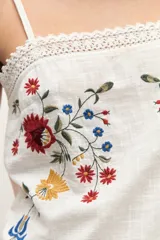 Top blanco de tirantes finos con detalle de encaje en escote y dobladillo, con bordado floral multicolor en el frente.